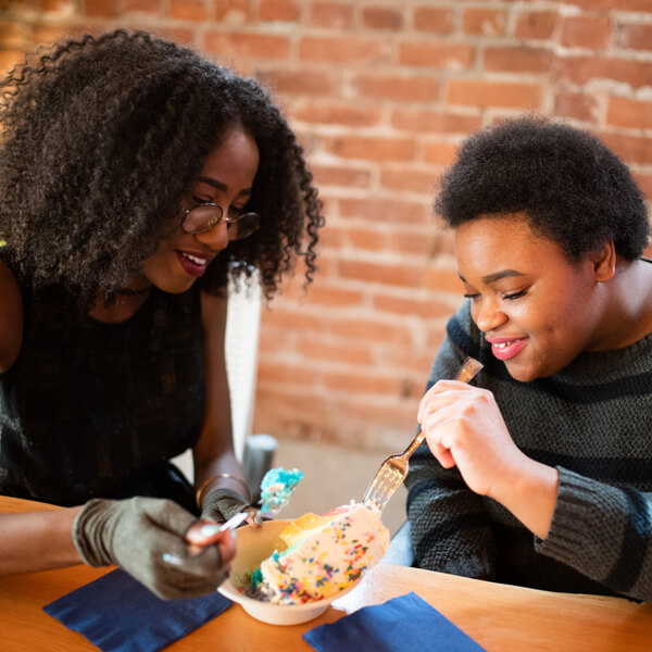 Two black people with feminine vibes are sharing a piece of cake with sprinkles on top. The person on the left has longer curly hair and glasses and the person on the right has short curly hair and no glasses. They are both smiling. Photo Credit: Disabled and Here
https://affecttheverb.com/collection/